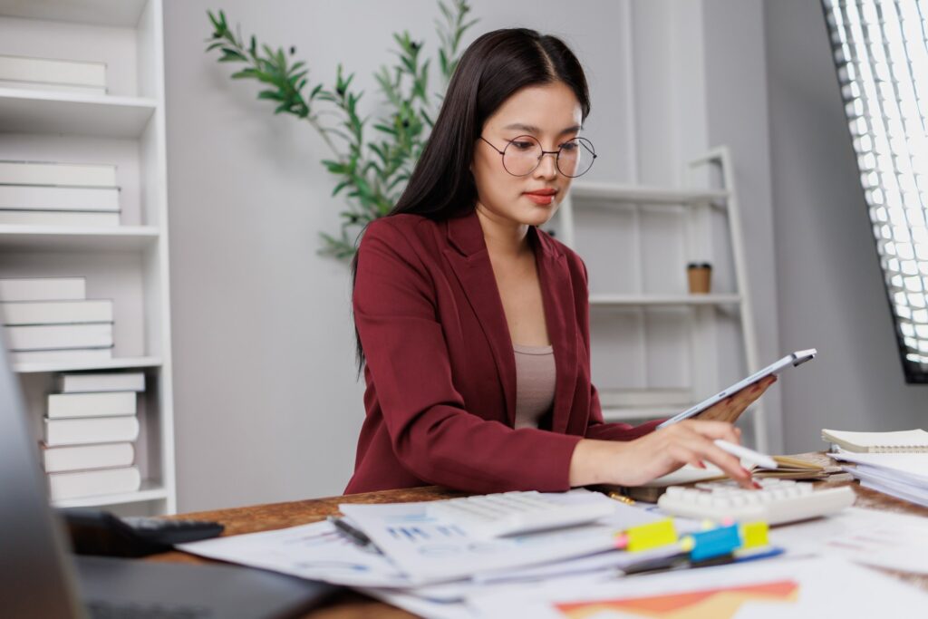 A woman in a burgundy blazer reviewing documents with a tablet and calculator at an office desk.