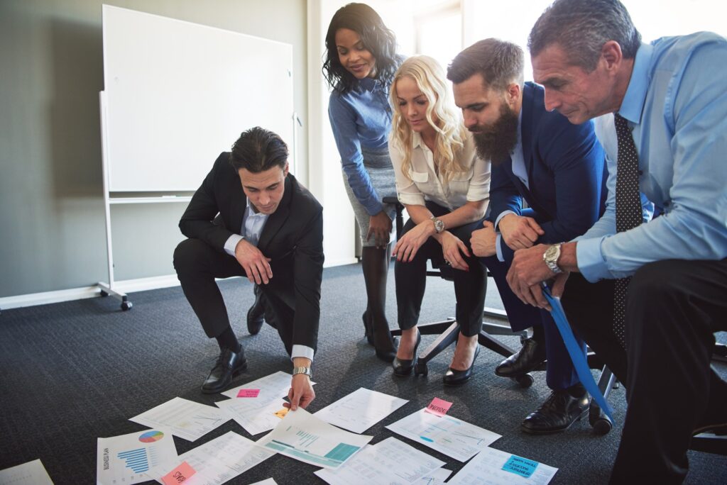 Five colleagues review charts spread across an office floor during a planning discussion.